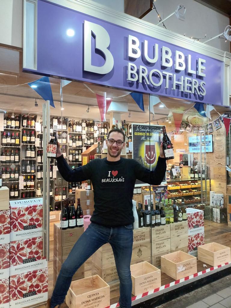 Rémi Matray standing outside Bubble Brothers' English Market shop in Cork, Ireland. He holds a bottle of Beaujolais wine in each hand.